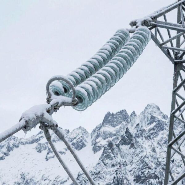 A close-up photograph captures icy power line insulators and hardware on a transmission tower against a backdrop of snow-covered mountains.