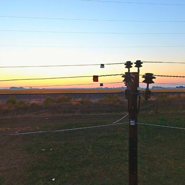 A wide shot of a rural landscape at sunset. A utility pole in the foreground has power lines extending from it, with small red and blue rectangular markers attached to the wires for identification or visibility.